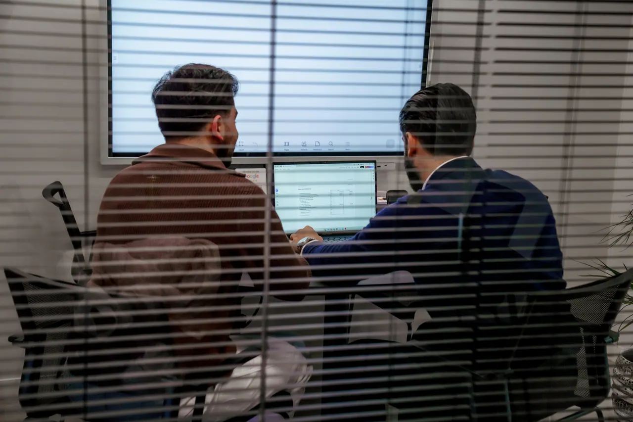 Twee collega's achter lamellen kijken samen naar een scherm, waar ze Make vs Zapier afwegen voor een flow